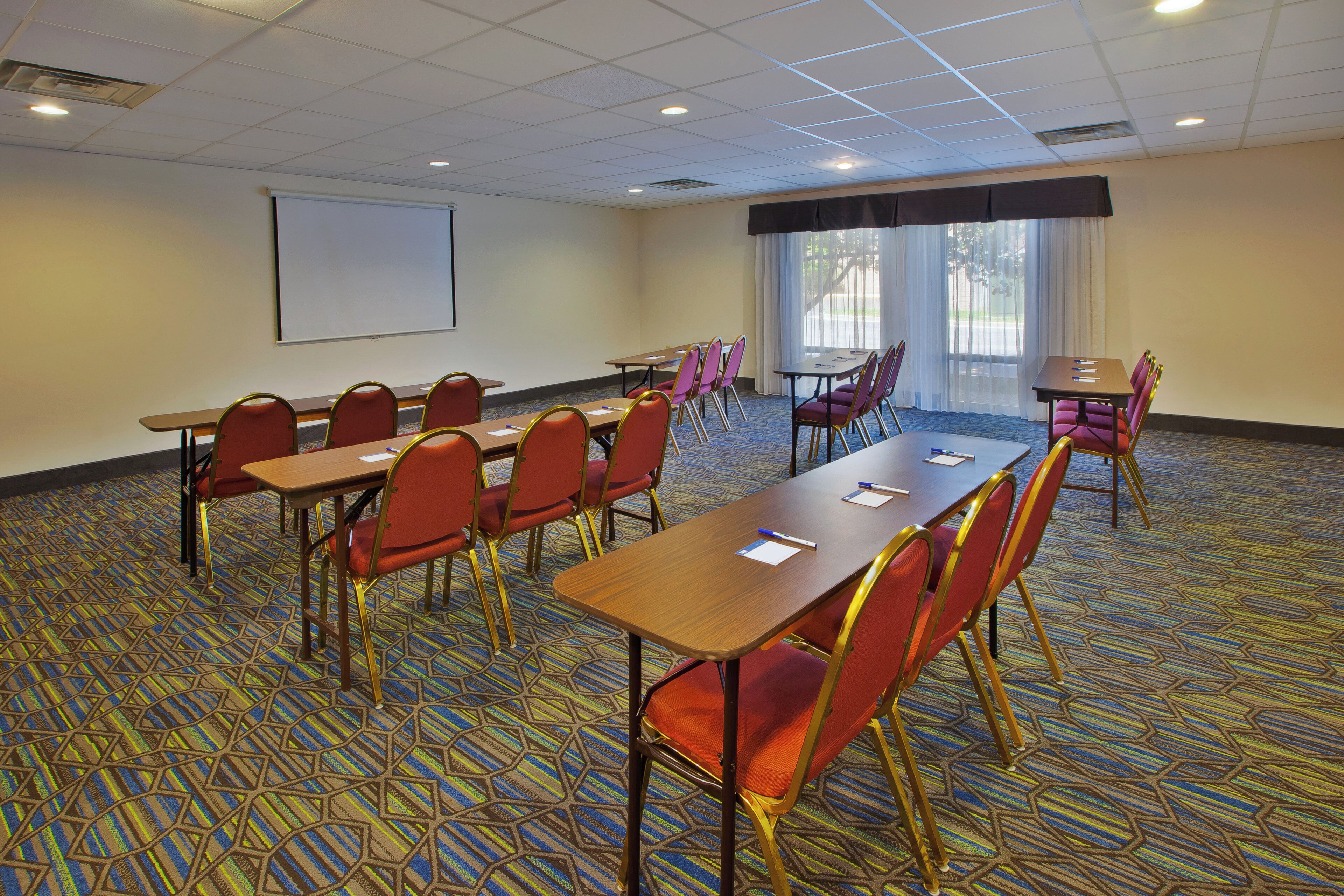 Un salón de conferencias con una larga mesa, varias sillas, una pantalla de proyector y un amplio ventanal con cortinas.