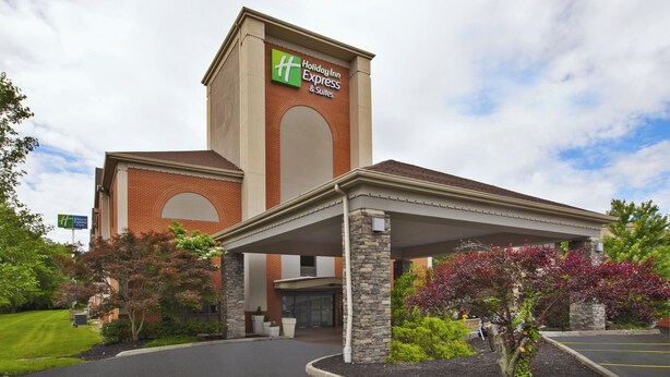 A Holiday Inn Express building with a brick facade, a covered entrance, and landscaped areas with shrubs and trees. - Holiday Inn Express & Suites Milford by IHG (Milford)