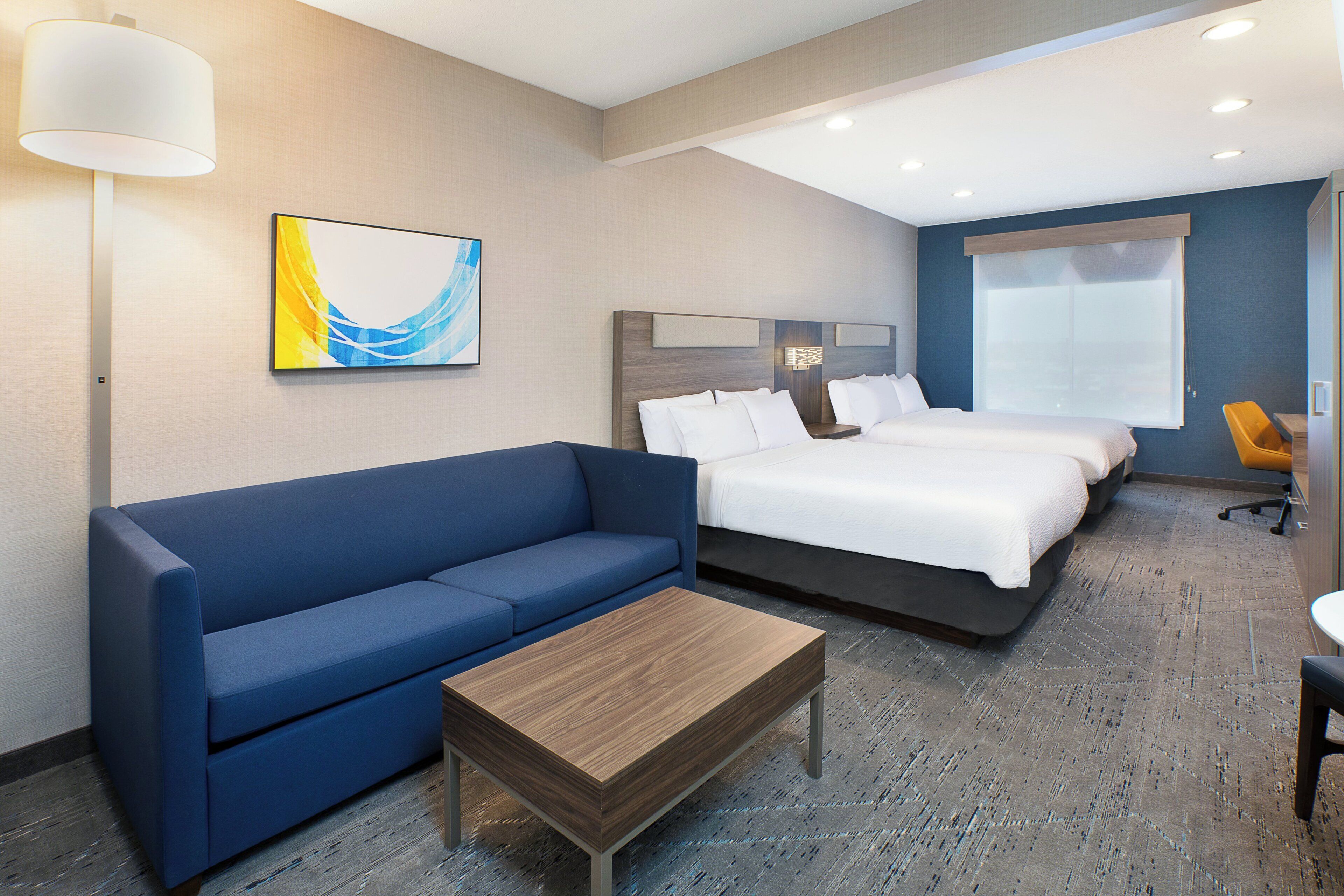 Habitación de hotel con un sofá azul, una mesa de centro de madera, dos camas con sábanas blancas, un cuadro en la pared y una ventana con persianas.
