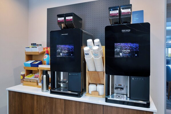 Two coffee machines with digital displays, a coffee dispenser, and a stack of disposable cups.