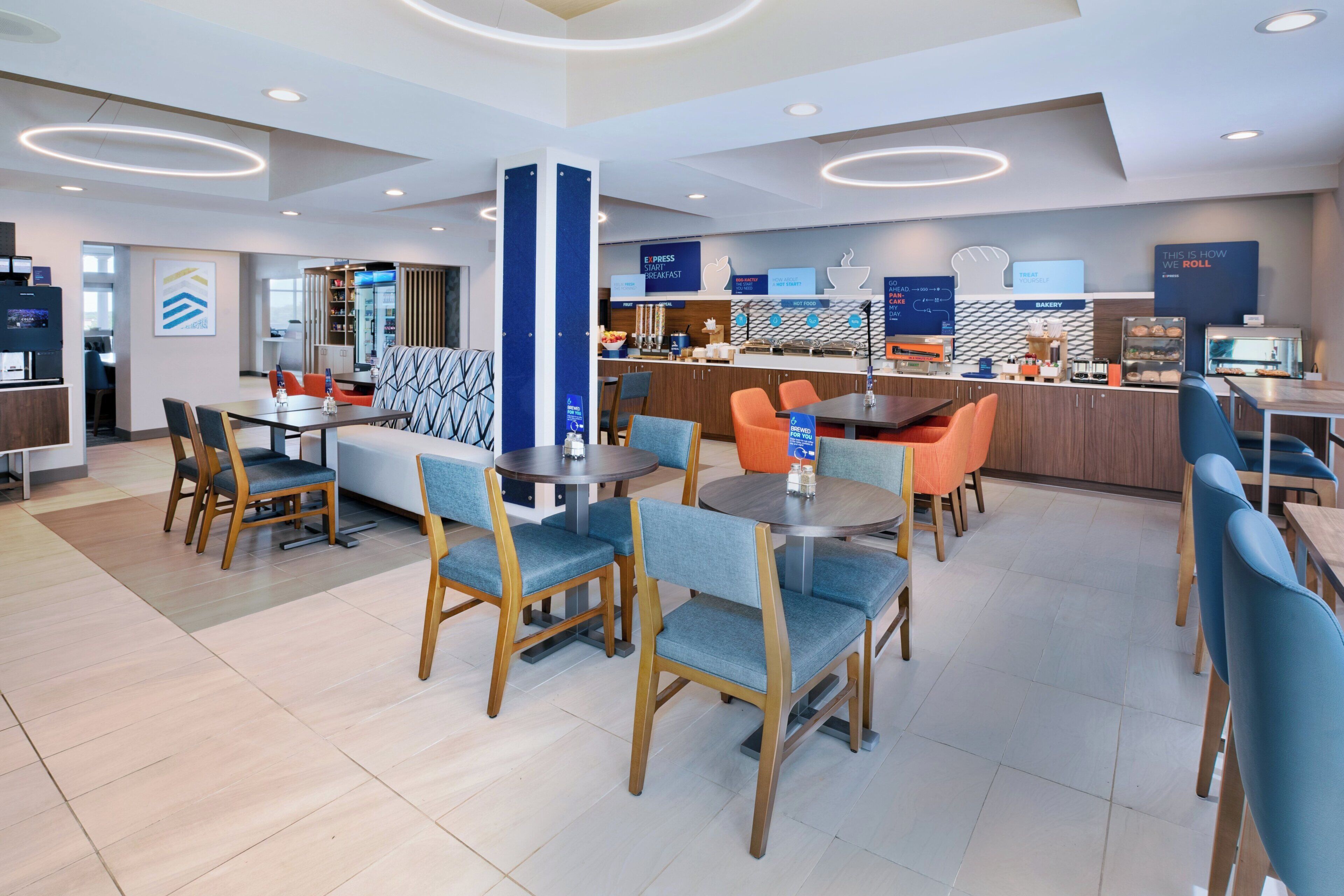 Restaurante de hotel moderno com várias áreas de estar, incluindo uma mesa de jantar central rodeada por cadeiras e uma área de estar separada com um sofá e cadeiras.