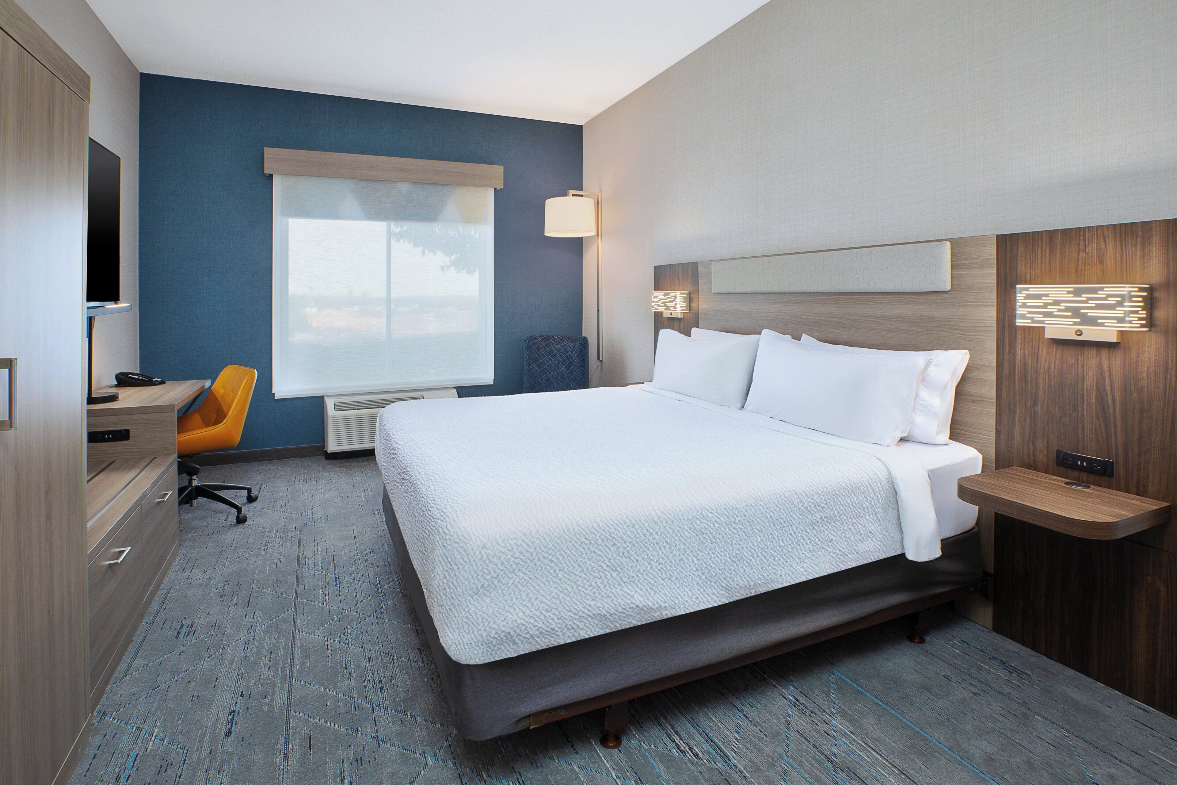 A hotel room with a large bed, a desk, a chair, a window with blinds, and a wall-mounted lamp.
