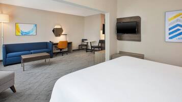 A hotel room with a large bed, a sofa, a desk with a chair, a TV mounted on the wall, and a painting on the wall.