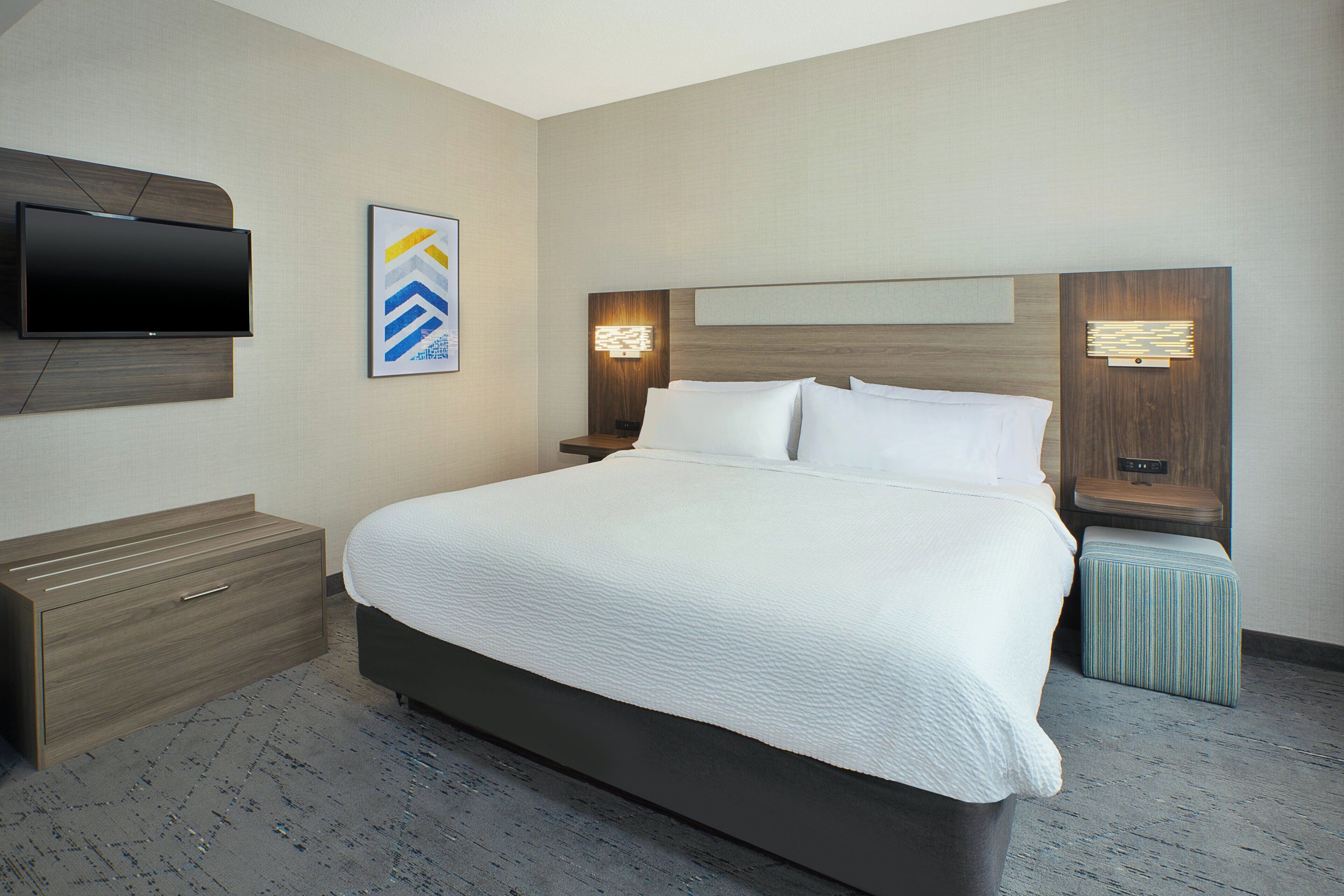 Habitación de hotel con una cama grande, un televisor de pantalla plana montado en la pared, una mesita de noche con una lámpara y un pequeño taburete.