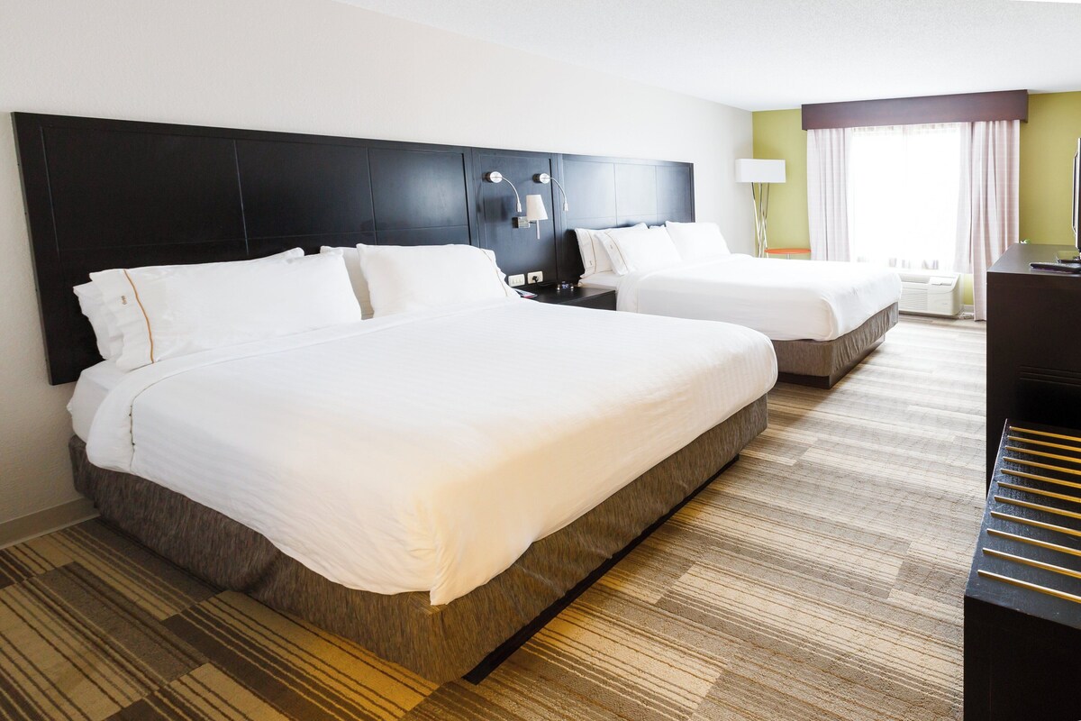 Une chambre d’hôtel avec deux lits, une tête de lit sombre, une lampe, un bureau et une grande fenêtre avec des rideaux.