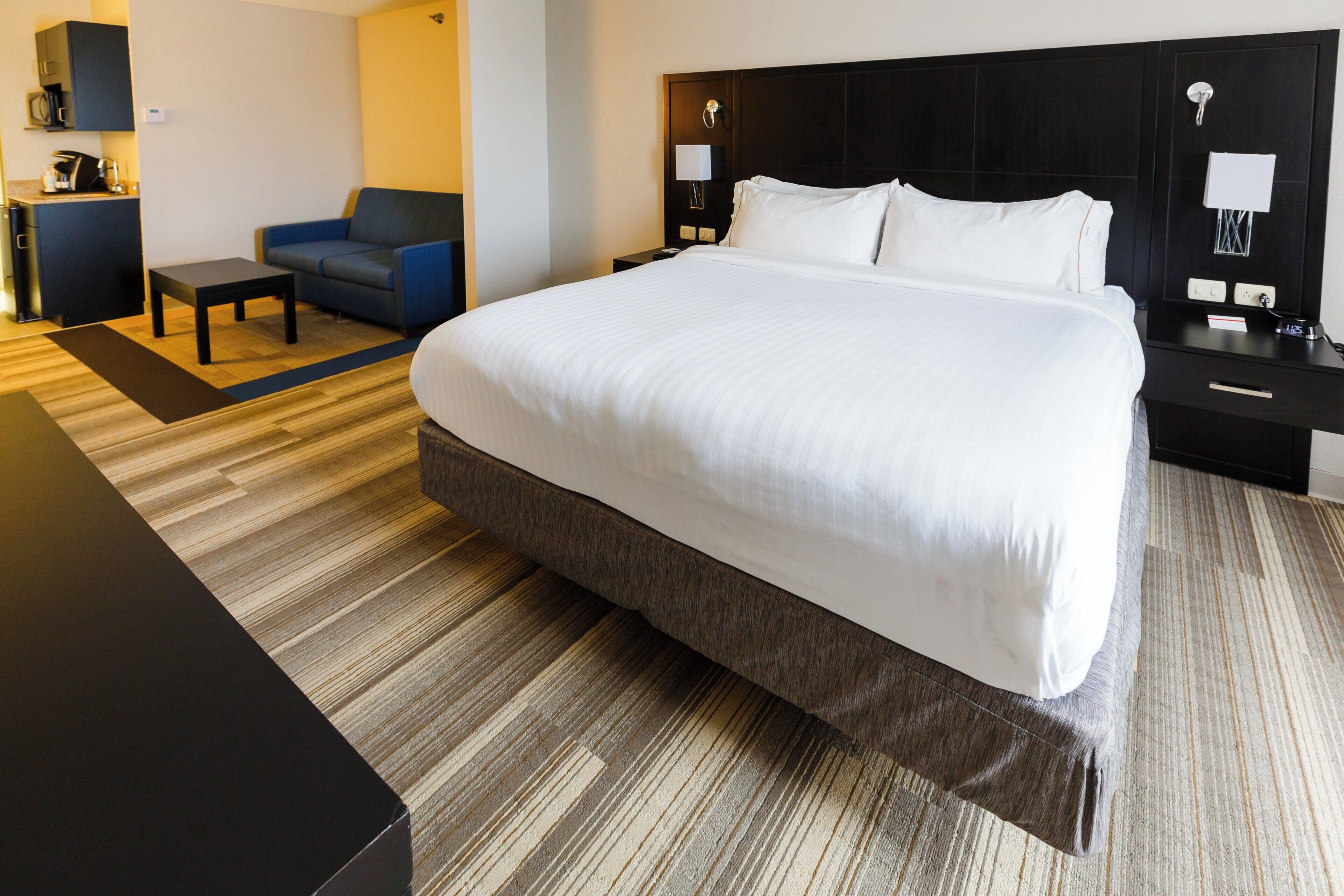 A hotel room with a large bed, a blue armchair, a small table, and a nightstand with a lamp.