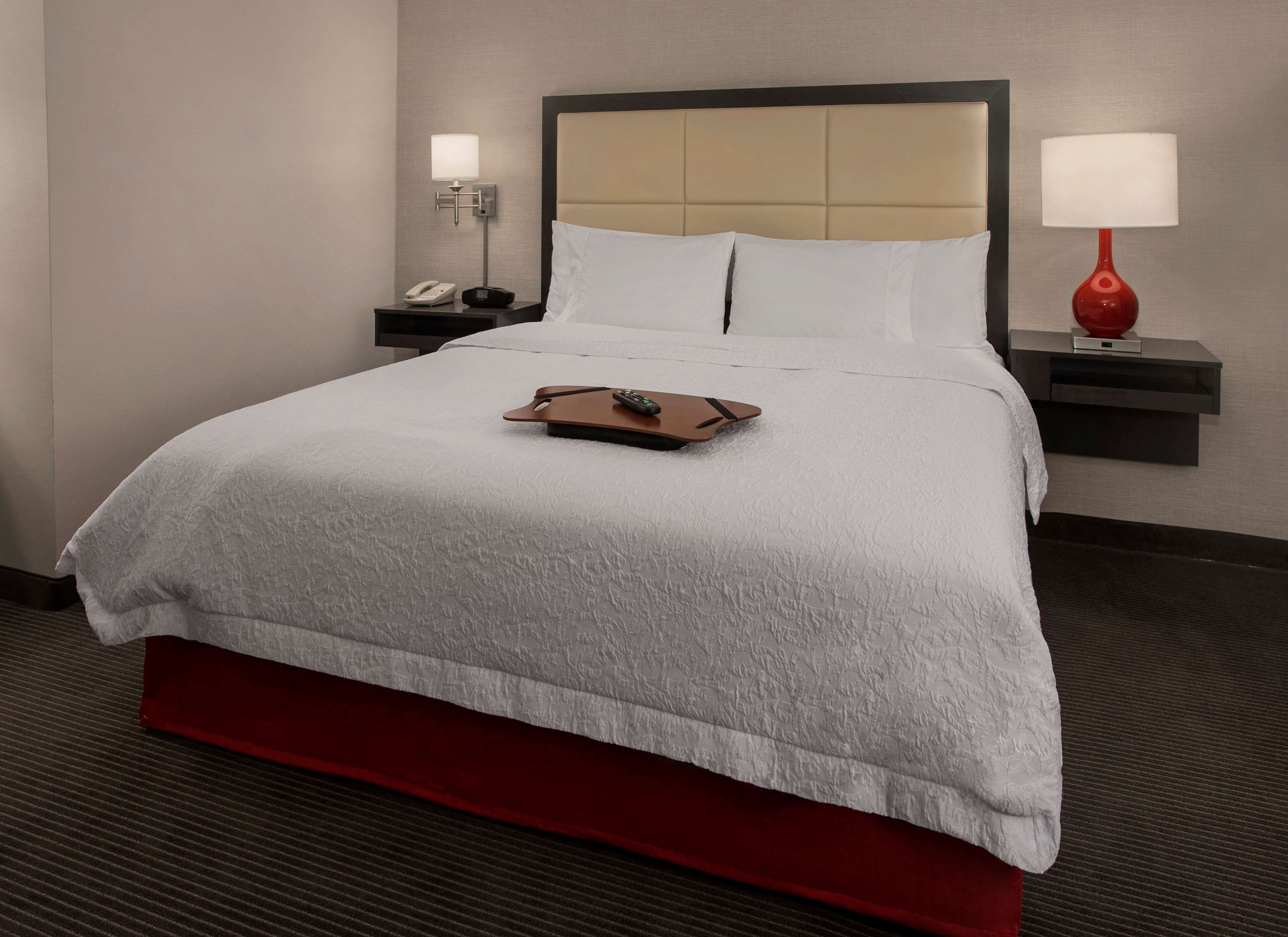 A hotel room with a large bed, a bedside table with a lamp, a phone, and a tray with a remote control.