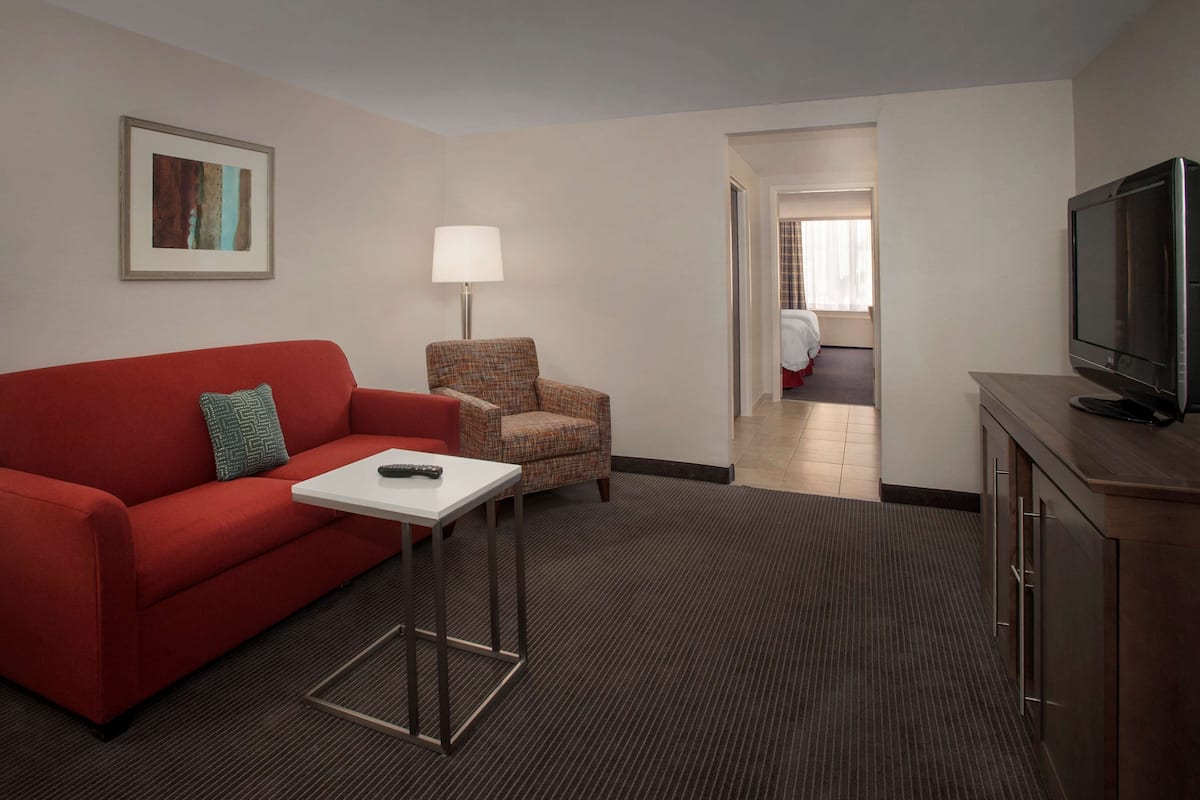 A hotel room with a red sofa, a plaid armchair, a small table with a remote control, a lamp, and a TV on a wooden cabinet.