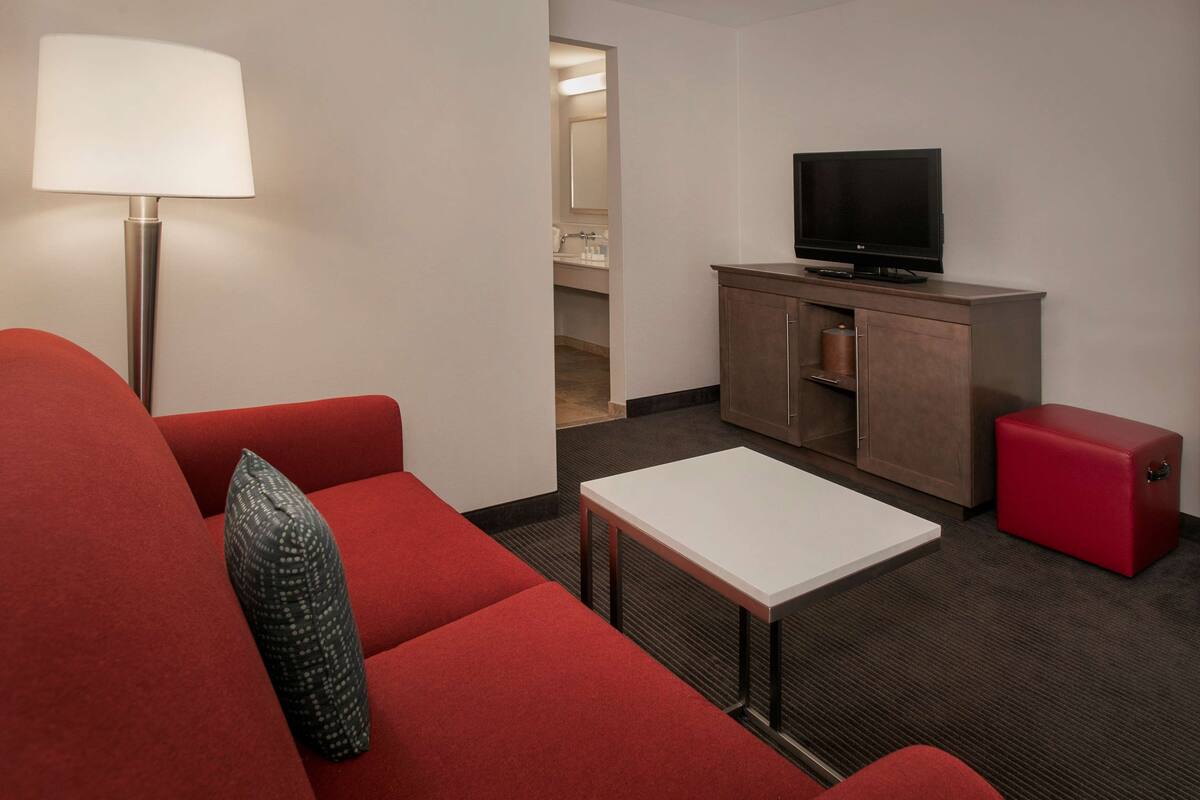 A compact hotel room with a red sofa, a small white table, a TV on a wooden cabinet, and a red storage unit.