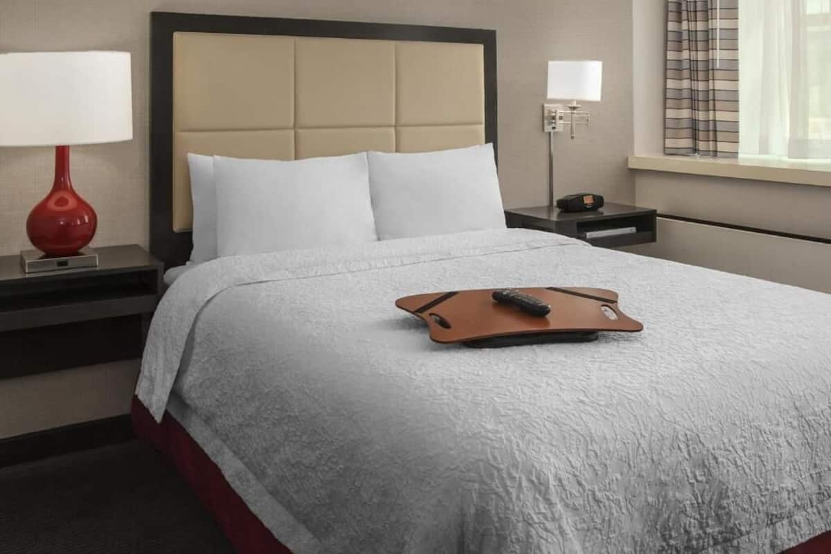 A hotel room with a bed, a nightstand, a red lamp, a remote control, and a window with curtains.