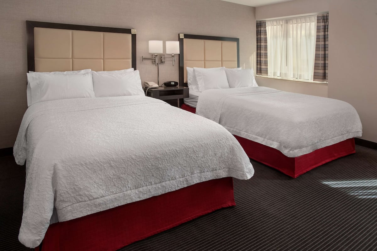 A hotel room with two beds, each with a red base and white bedding. There is a nightstand with a lamp and a phone between the beds.
