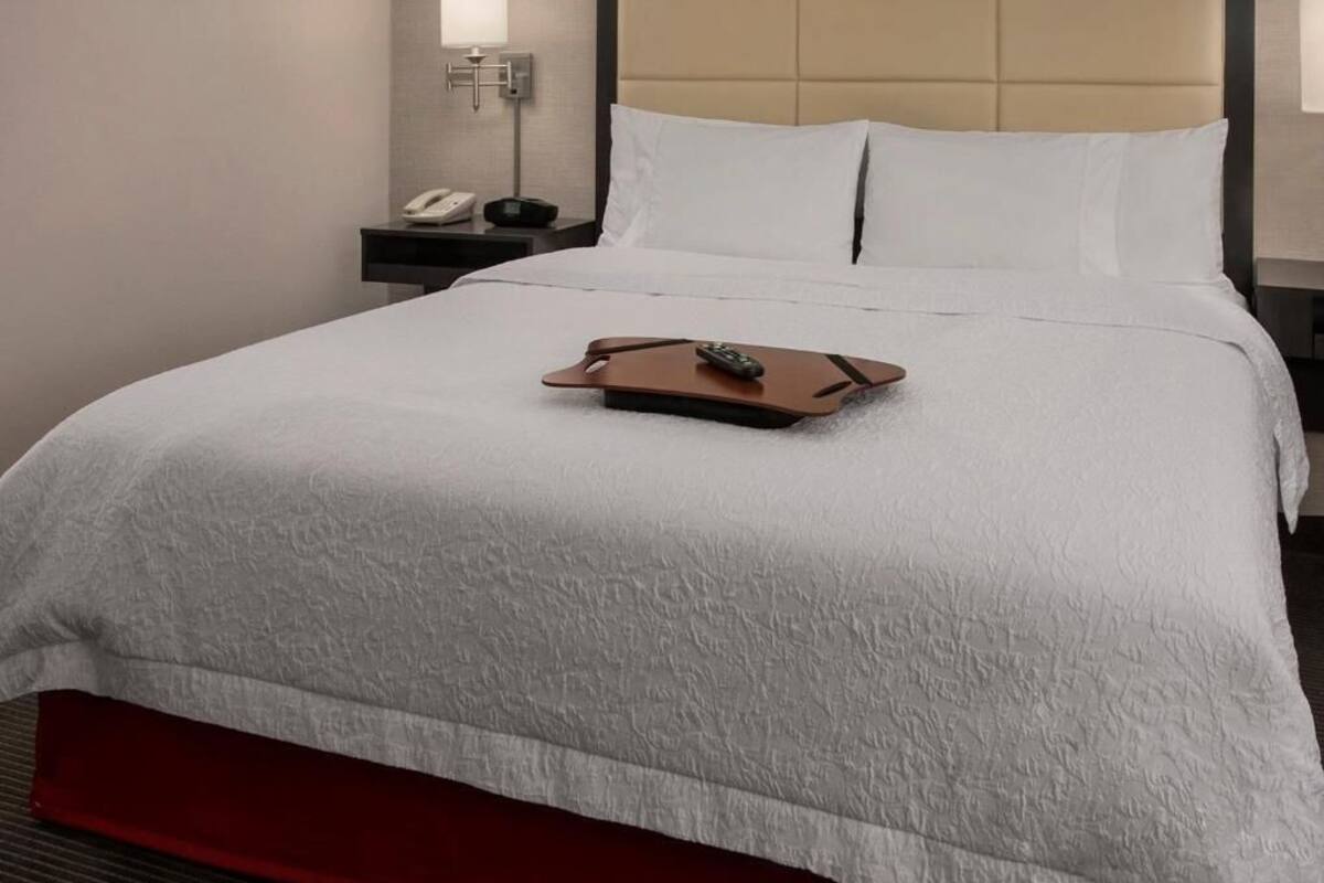 A neatly made bed with a remote control on a tray, a bedside phone, and a lamp on a nightstand.