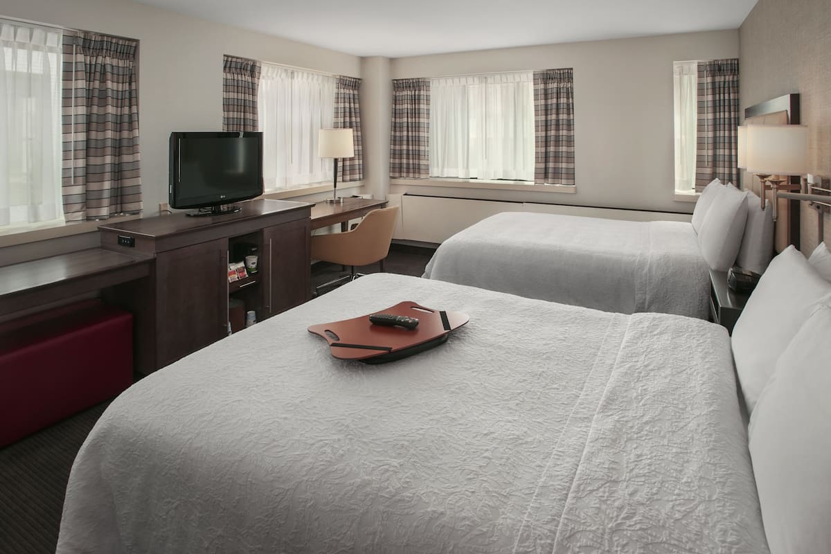 A hotel room with two beds, a desk with a television, a chair, and a red bench.