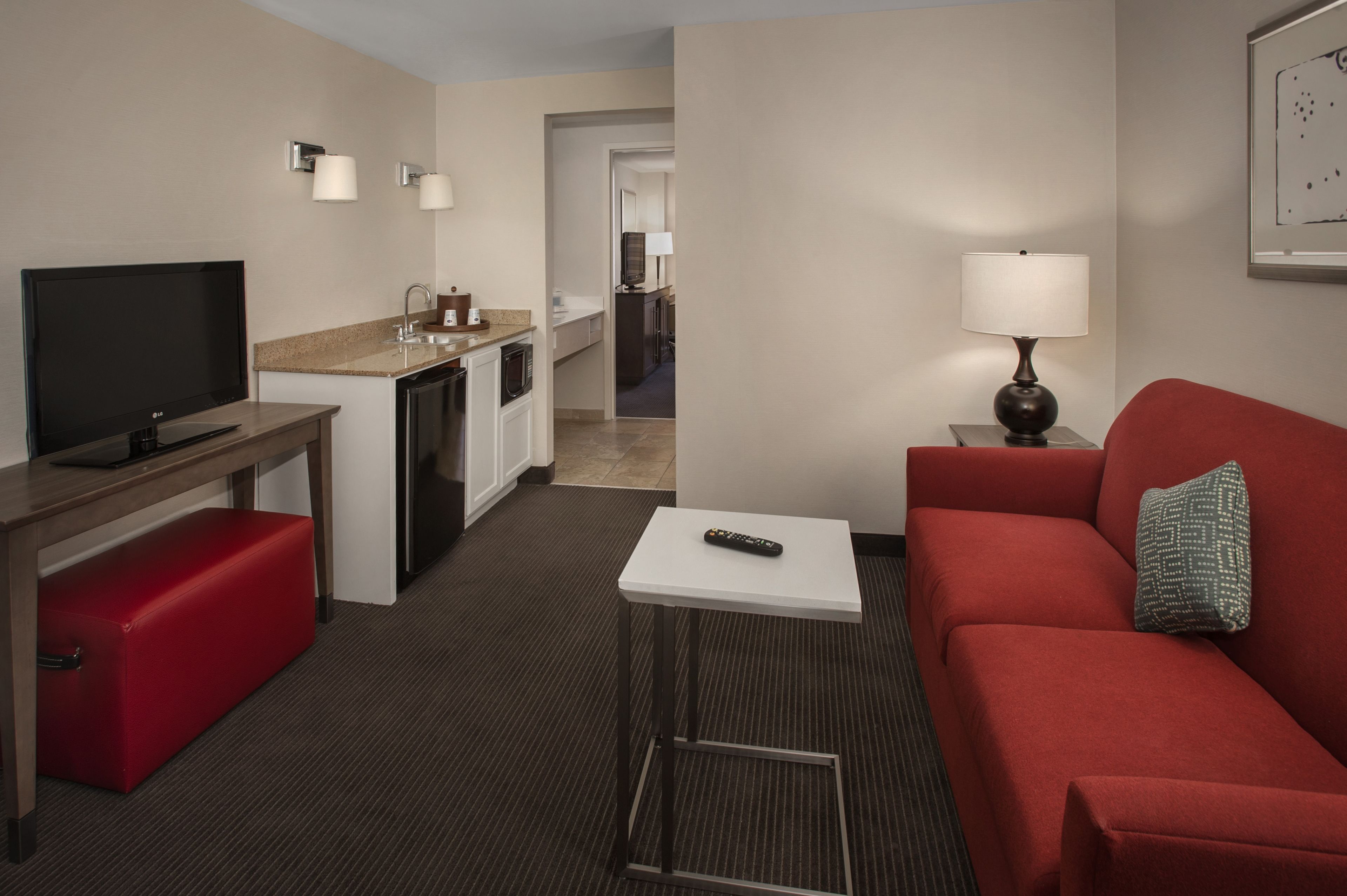 A hotel room with a TV, a red sofa, a small table with a remote, a lamp, and a kitchenette.