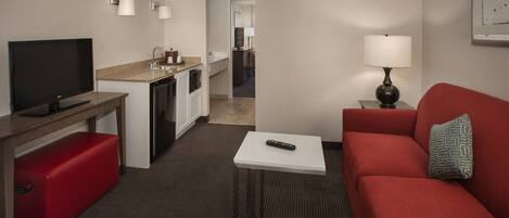 A hotel room with a TV, a red sofa, a small table with a remote, a lamp, and a kitchenette.
