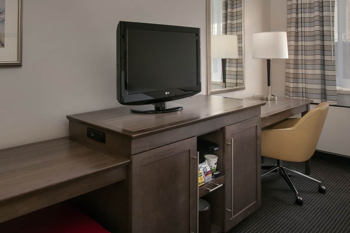 A hotel room with a wooden desk, a television, a mirror, a lamp, a chair, and a small cabinet with a trash bin.