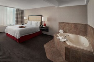 Una habitación de hotel con cama, mesita de noche, lámpara, baño con bañera y ventana con cortinas.