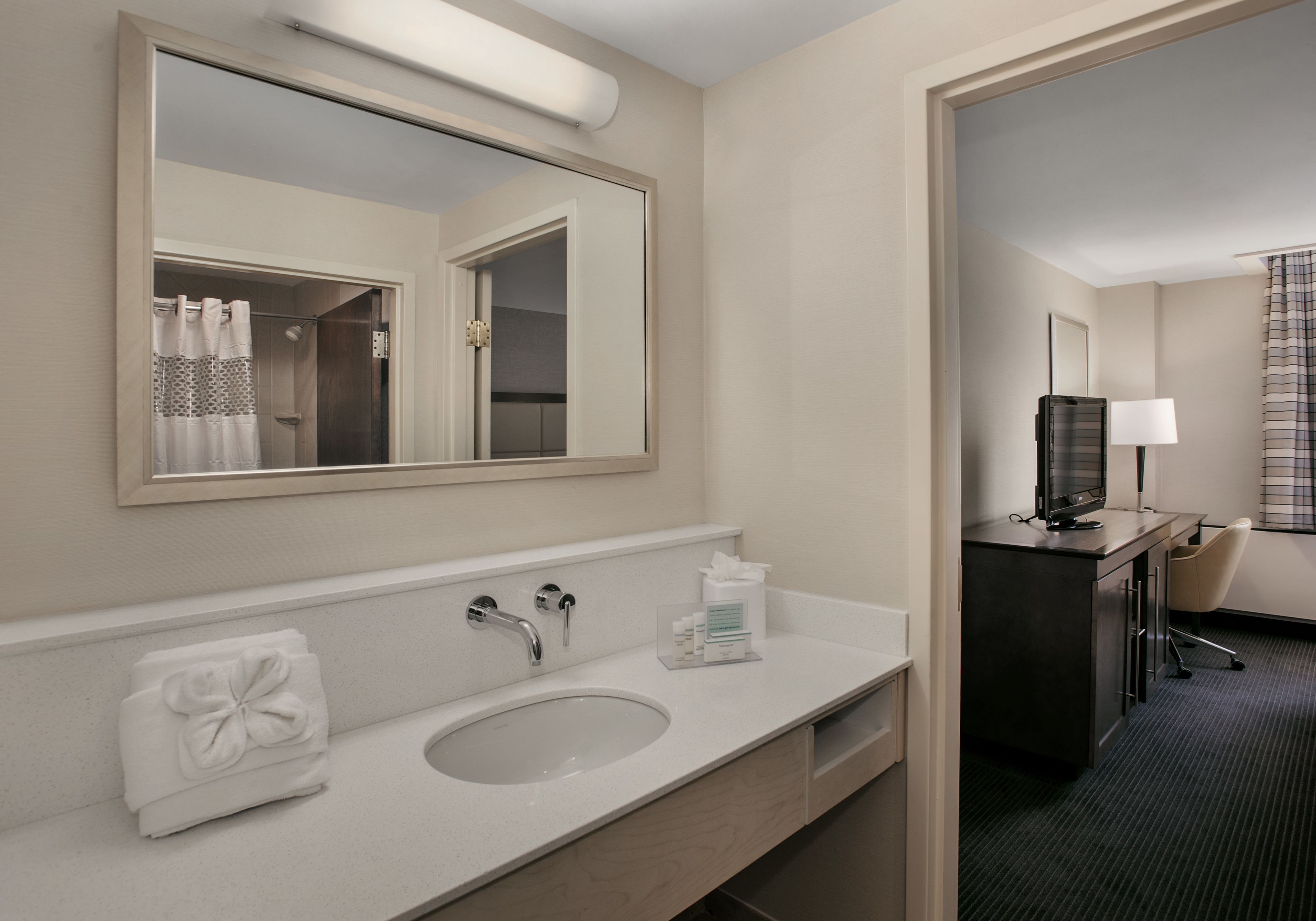 Une salle de bain d’hôtel équipée d’un lavabo, d’un miroir, d’une serviette, d’un bureau avec une lampe et un téléviseur.