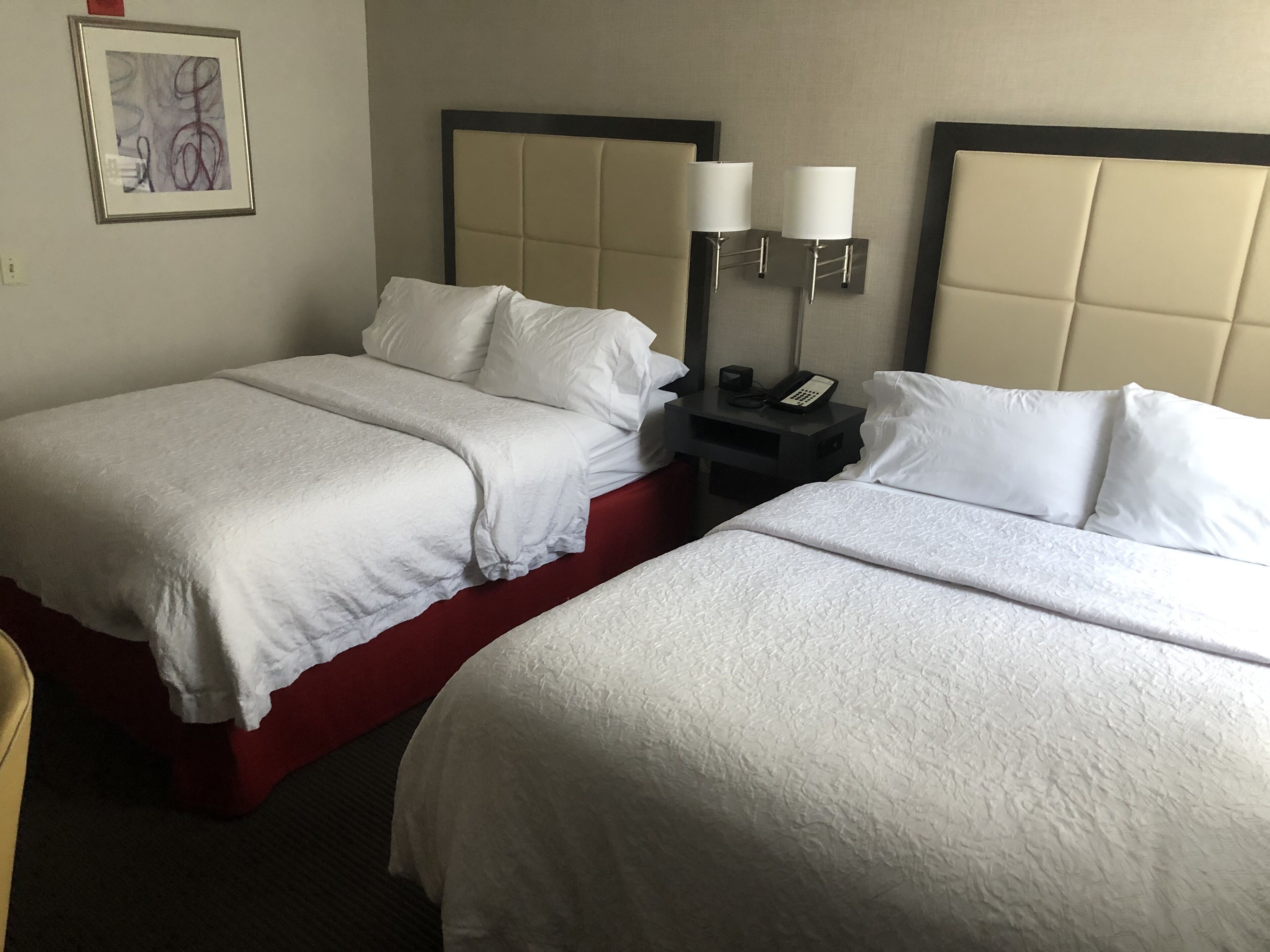 A hotel room with two beds, each with white bedding and red headboards. There is a nightstand between the beds with two wall-mounted lamps and a phone.