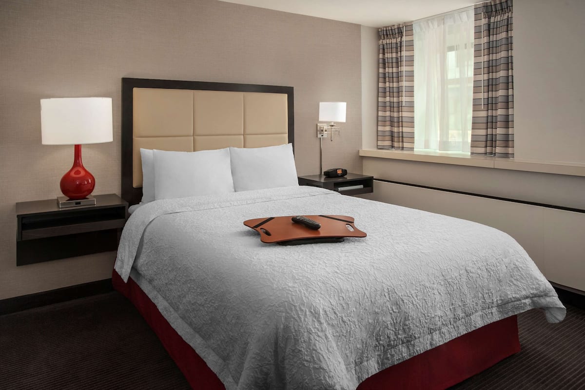 A hotel room with a bed, two bedside tables, a lamp, a remote control, and a window with curtains.