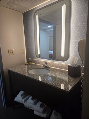 A modern bathroom with a granite sink, a large mirror with integrated lighting, a towel rack, and a tissue dispenser.