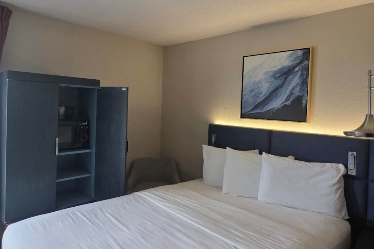 A hotel room with a bed, a desk with a chair, a wardrobe, and a painting on the wall.