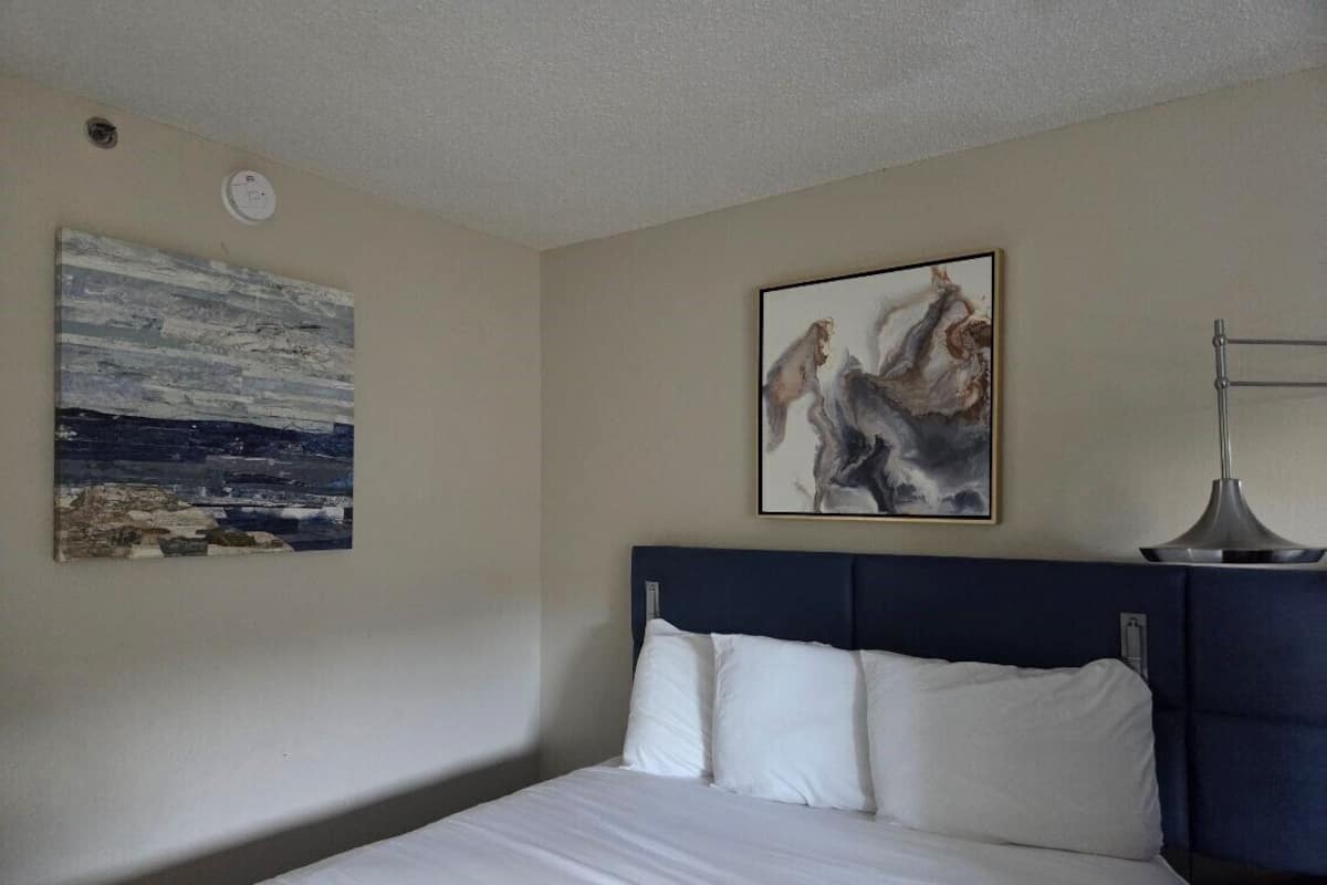 A hotel room with a bed, a headboard, a wall-mounted lamp, and two pieces of wall art.
