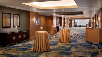 A hotel lobby with a high ceiling, large windows, and a patterned carpet. There are round tables with tablecloths and a reception desk.