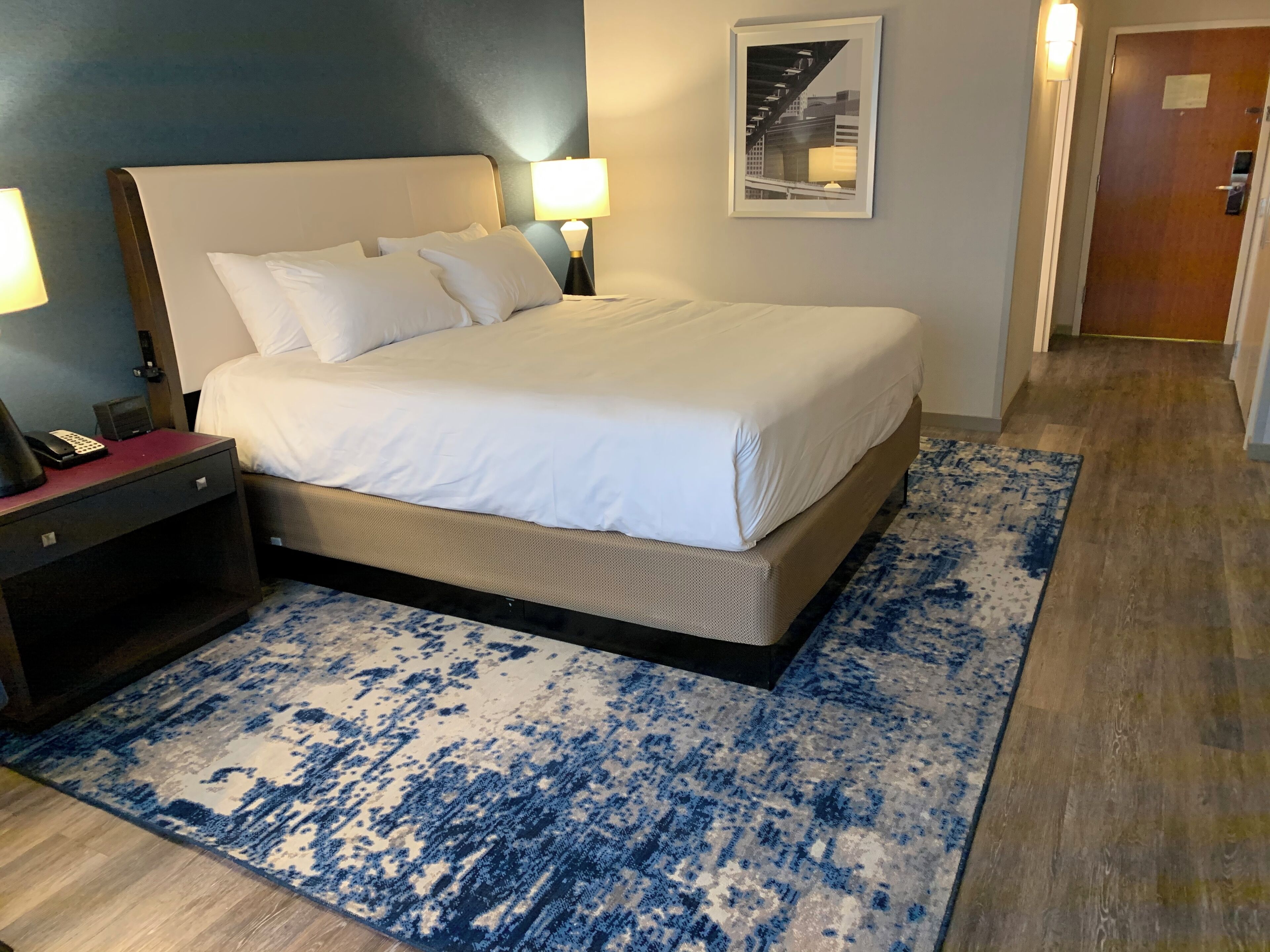 A neatly made bed with white linens, a blue and white patterned rug, a nightstand with a lamp, and a framed picture on the wall.