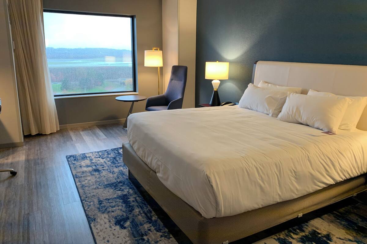 A hotel room with a large bed, a chair, a small table, and a window with a view.