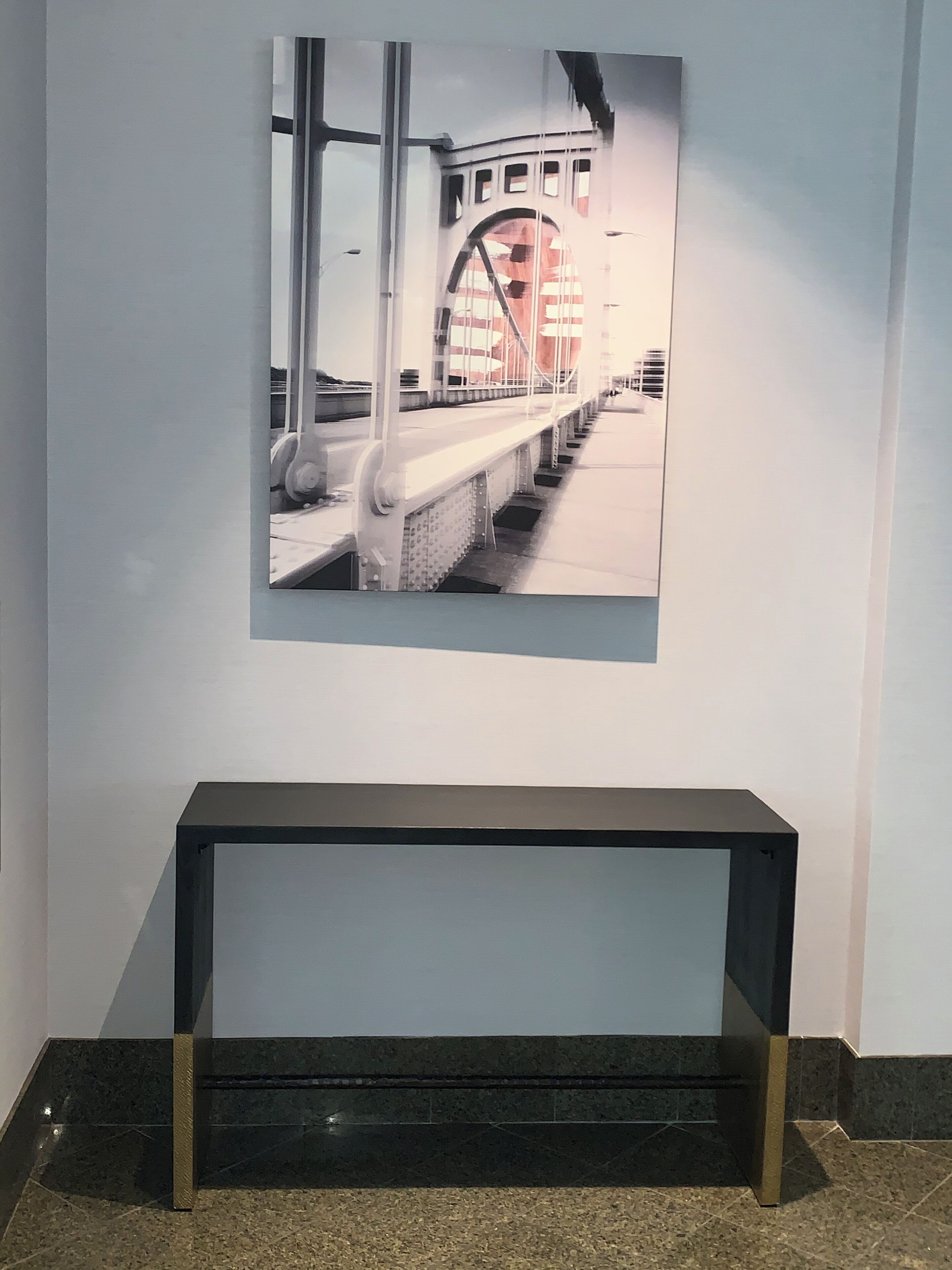 Fotografía enmarco de un puente en una pared, un banco negro al frente y un piso de baldosas.