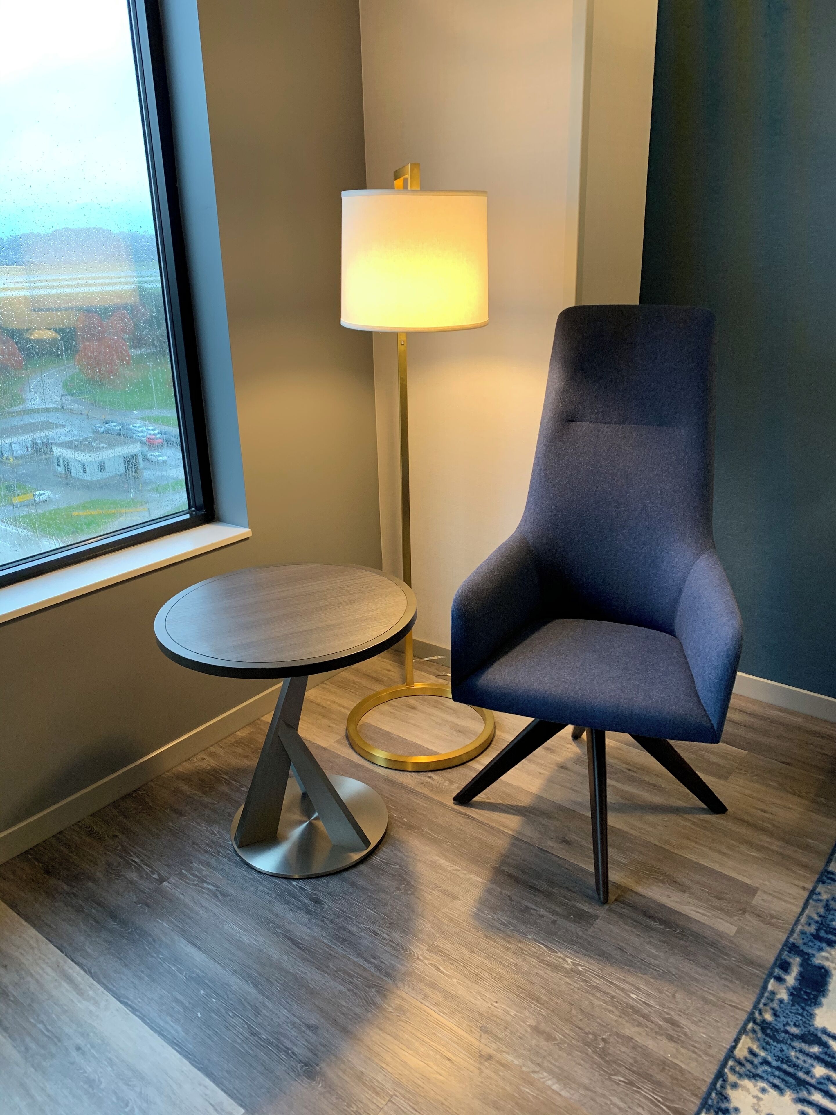 A modern room with a round table, a blue armchair, a floor lamp, and a view of a parking lot through a window.