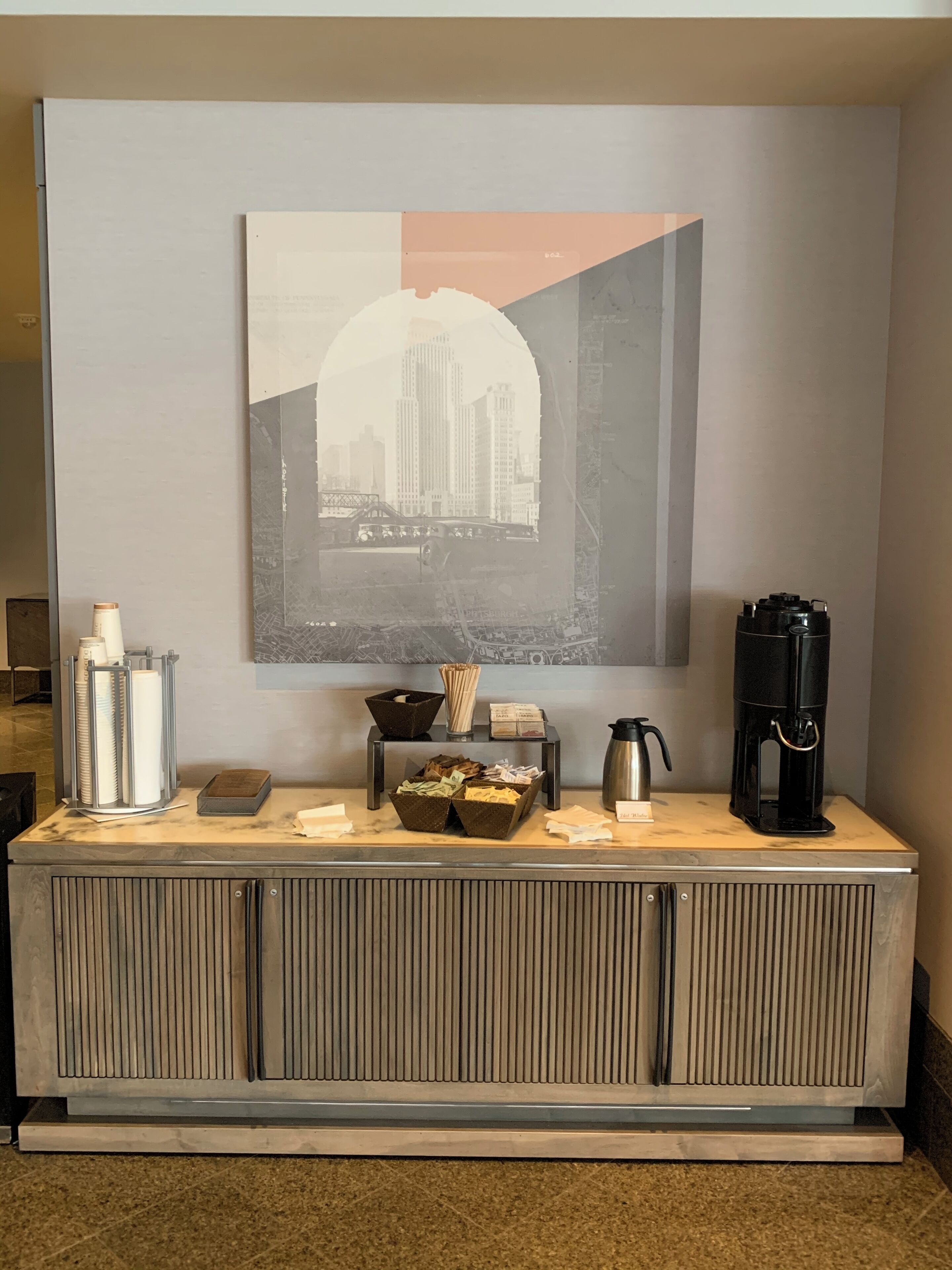 A hotel reception area with a marble counter, a coffee maker, a kettle, and a framed cityscape picture above.