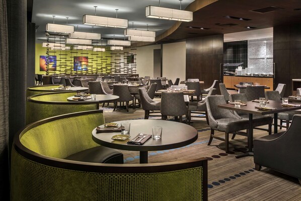 A modern restaurant with round tables and green upholstered booths, a kitchen in the background, and pendant lighting.