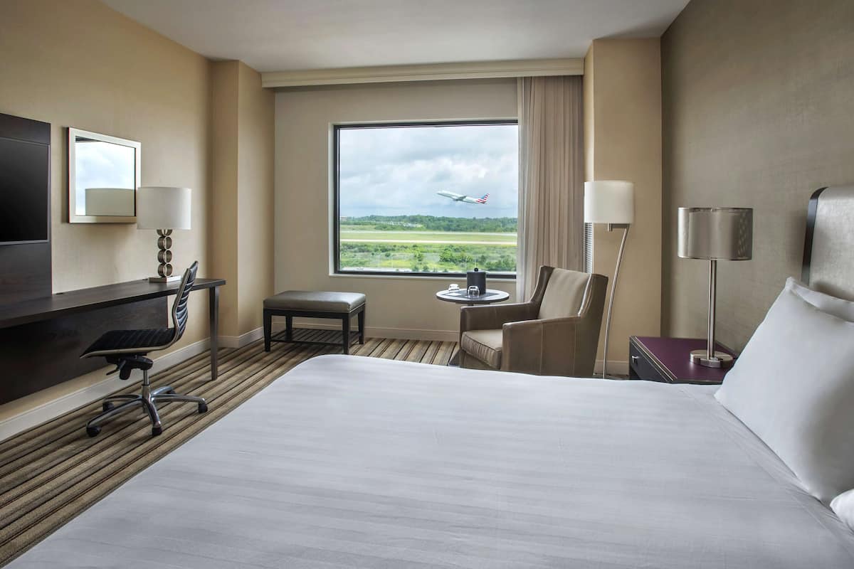 A hotel room with a large bed, a desk with a chair, a small table with a chair, a TV, and a window with a view of an airport.
