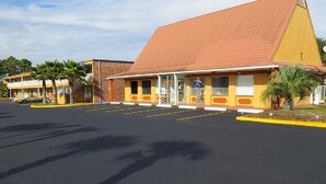A motel with a red roof and yellow exterior walls, featuring a parking lot with marked spaces and palm trees.