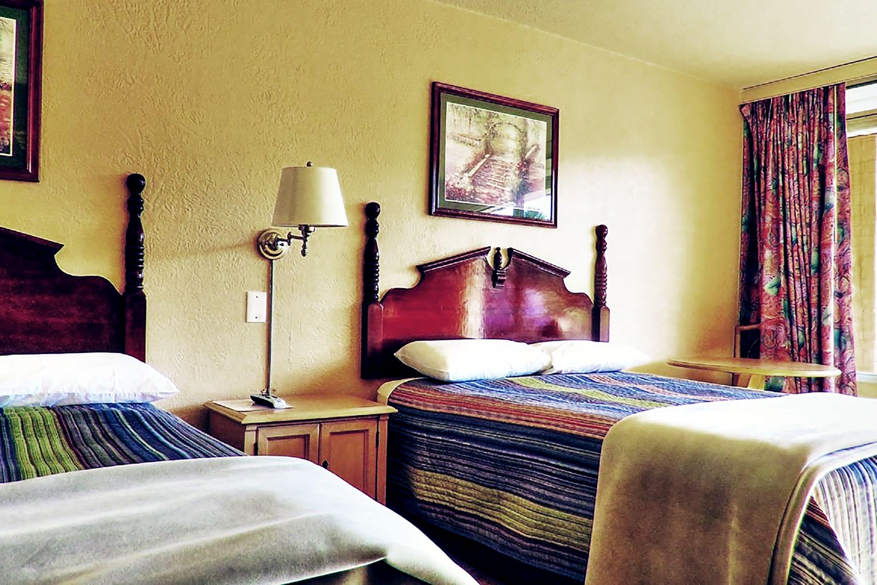A hotel room with two beds, a nightstand with a lamp, a framed picture on the wall, and a window with curtains.