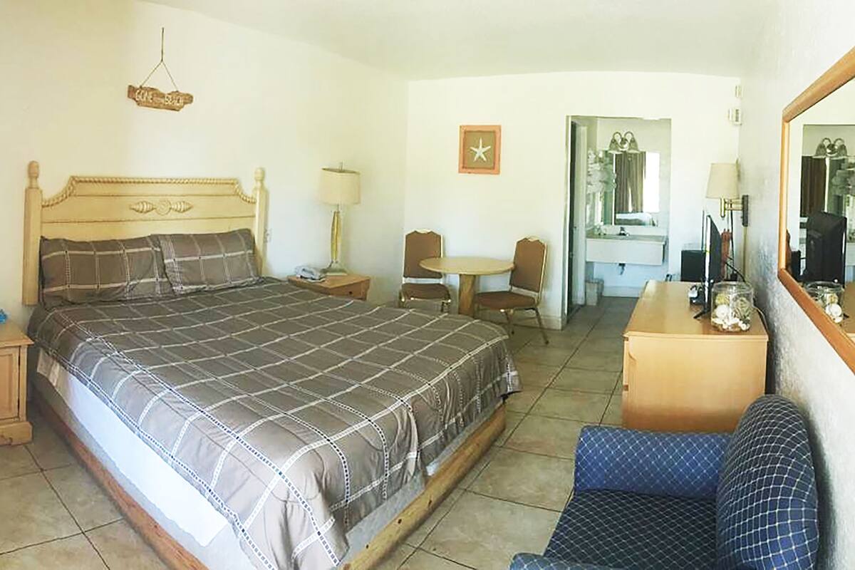 A bedroom with a large bed, a desk with a chair, a small table, a mirror, and a hanging sign.