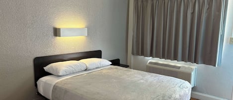 Una cama bien hecha con sábanas blancas, una lámpara de pared, una ventana con cortinas y una rejilla de ventilación en el techo.