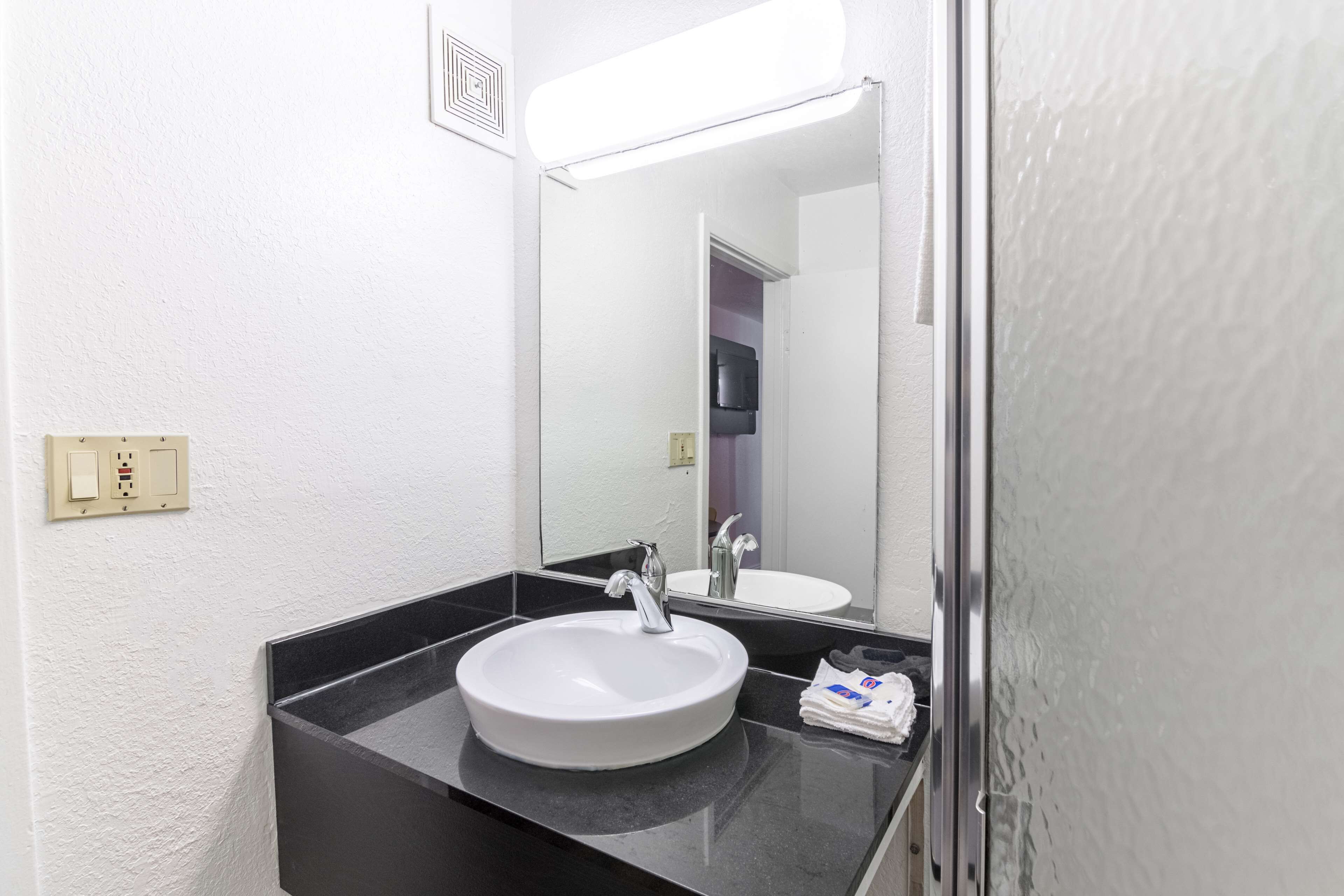 A bathroom with a white sink, black countertop, and a mirror reflecting a door and a TV.