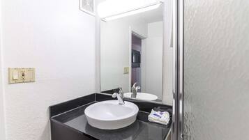 Une salle de bain avec un lavabo blanc, un plan de toilette noir et un miroir qui reflète une porte et un téléviseur.