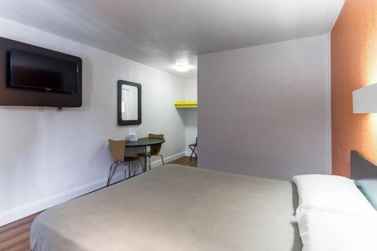 A hotel room with a bed, a TV mounted on the wall, a small table with chairs, and a yellow shelf.