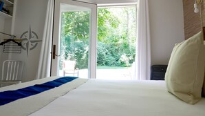 Un dormitorio con cama, una silla blanca, una estantería con libros, una ventana con cortinas y vistas al exterior con zonas verdes.
