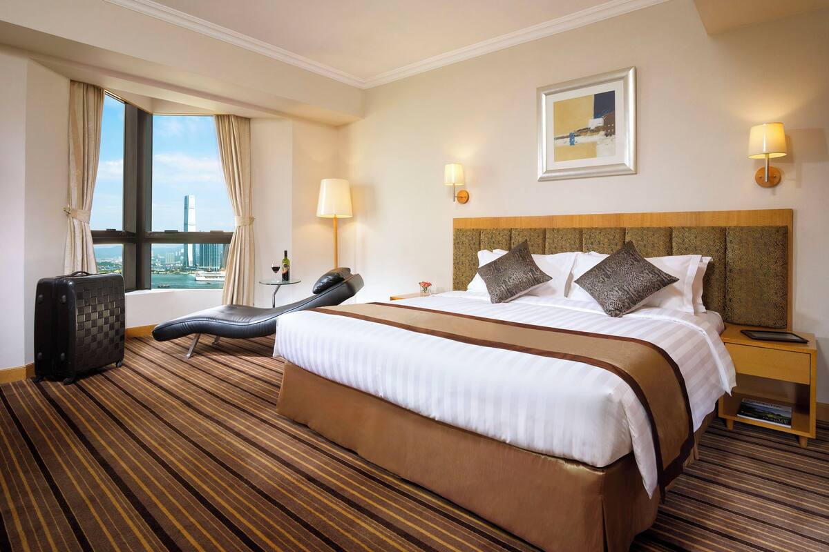 Quarto de hotel com uma cama grande, área de estar com uma poltrona, mala, abajur e vista para a paisagem urbana.
