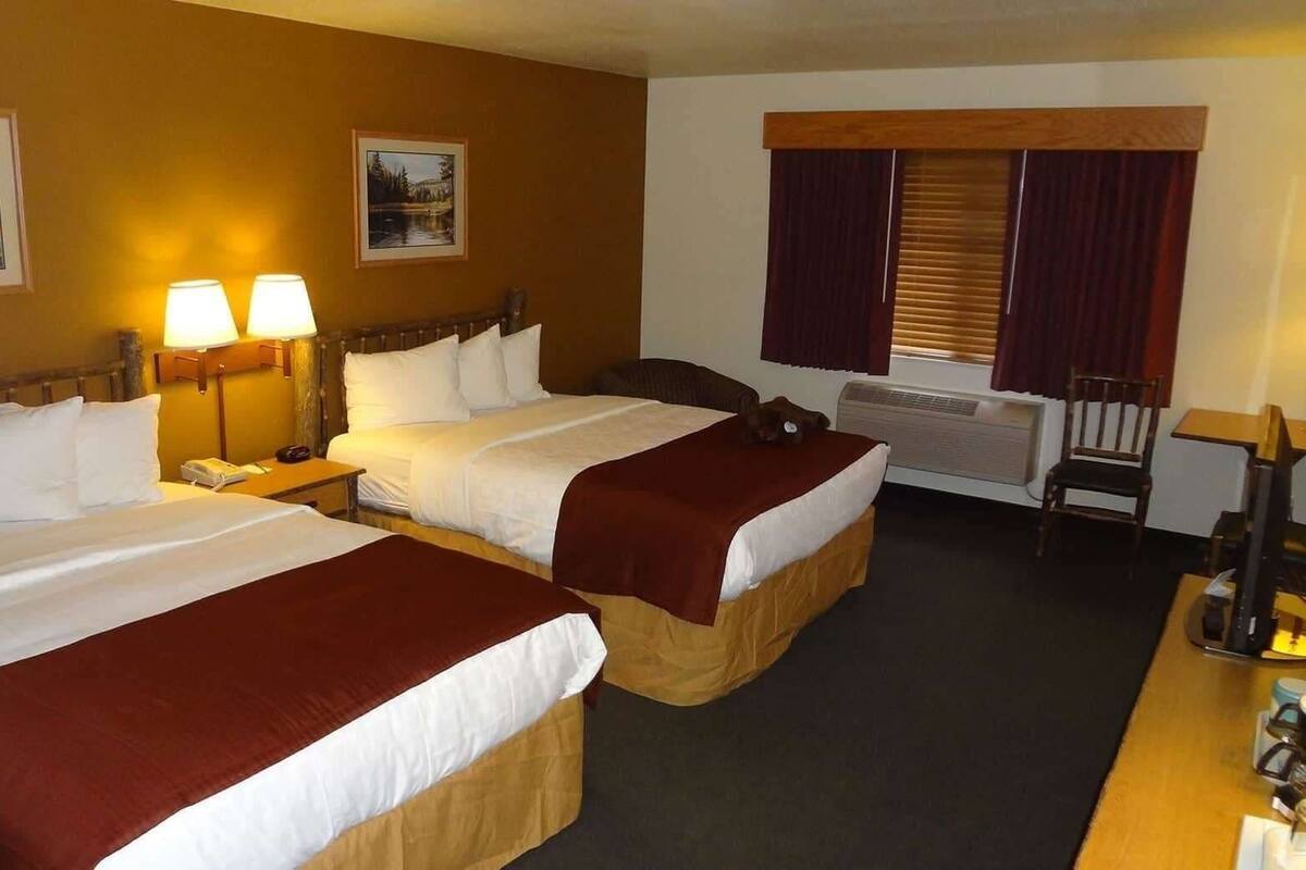 A hotel room with two beds, a desk, a chair, a window with blinds, and a wall-mounted heater.