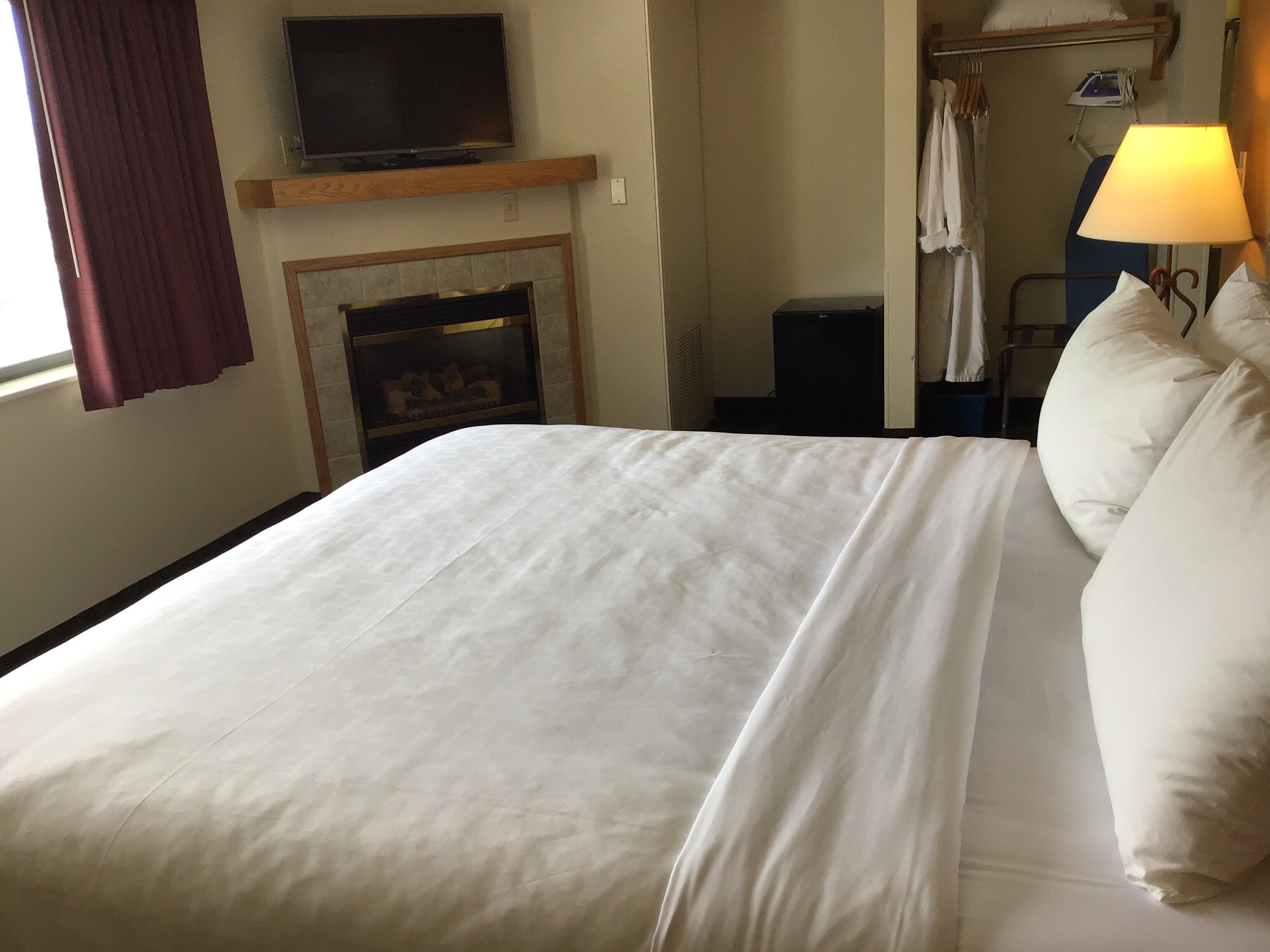 A hotel room with a large bed, a television on a wooden shelf above a fireplace, a lamp, and a wardrobe.