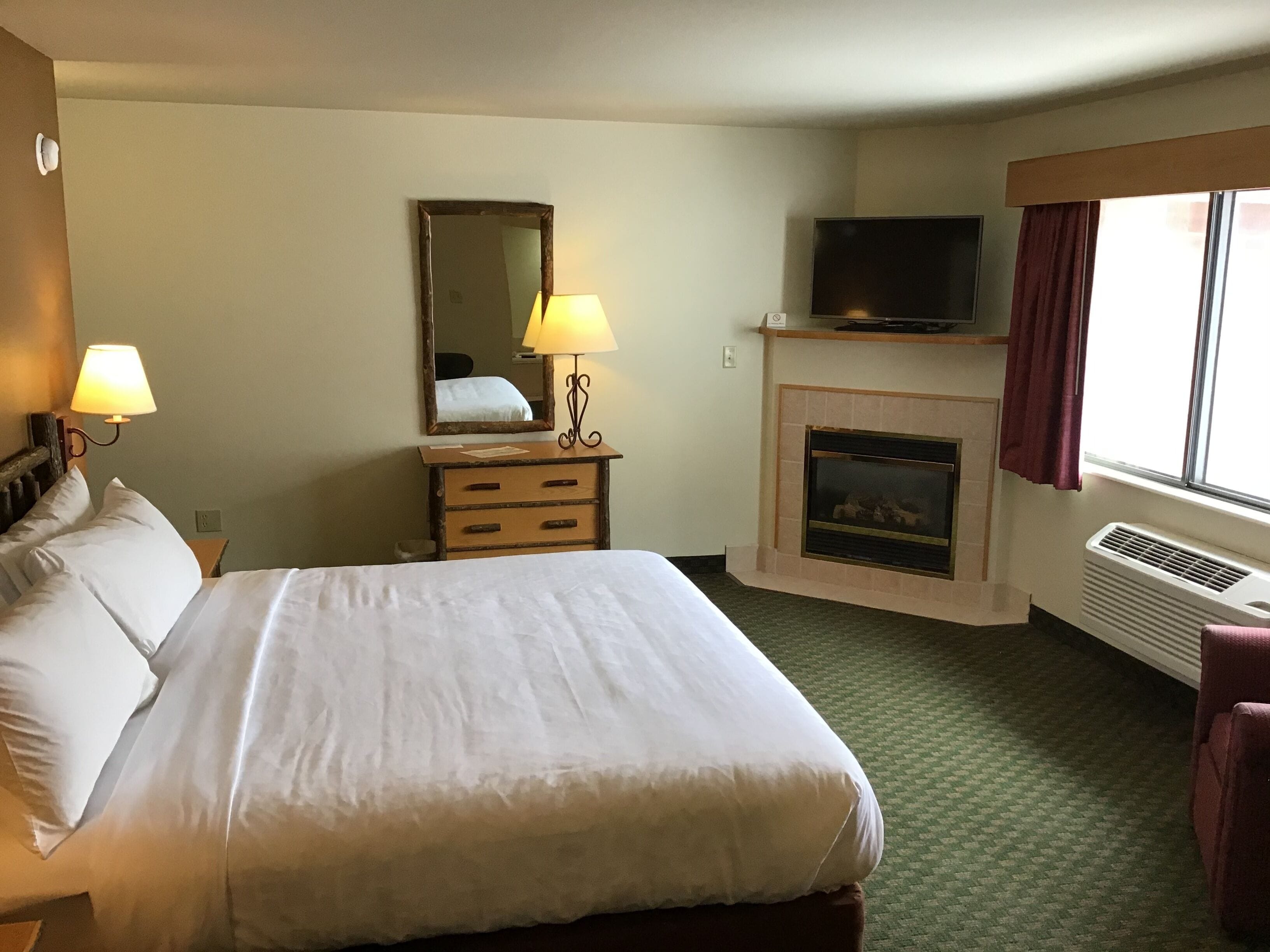 A hotel room with a bed, a dresser, a mirror, a television, a fireplace, and a window with curtains.