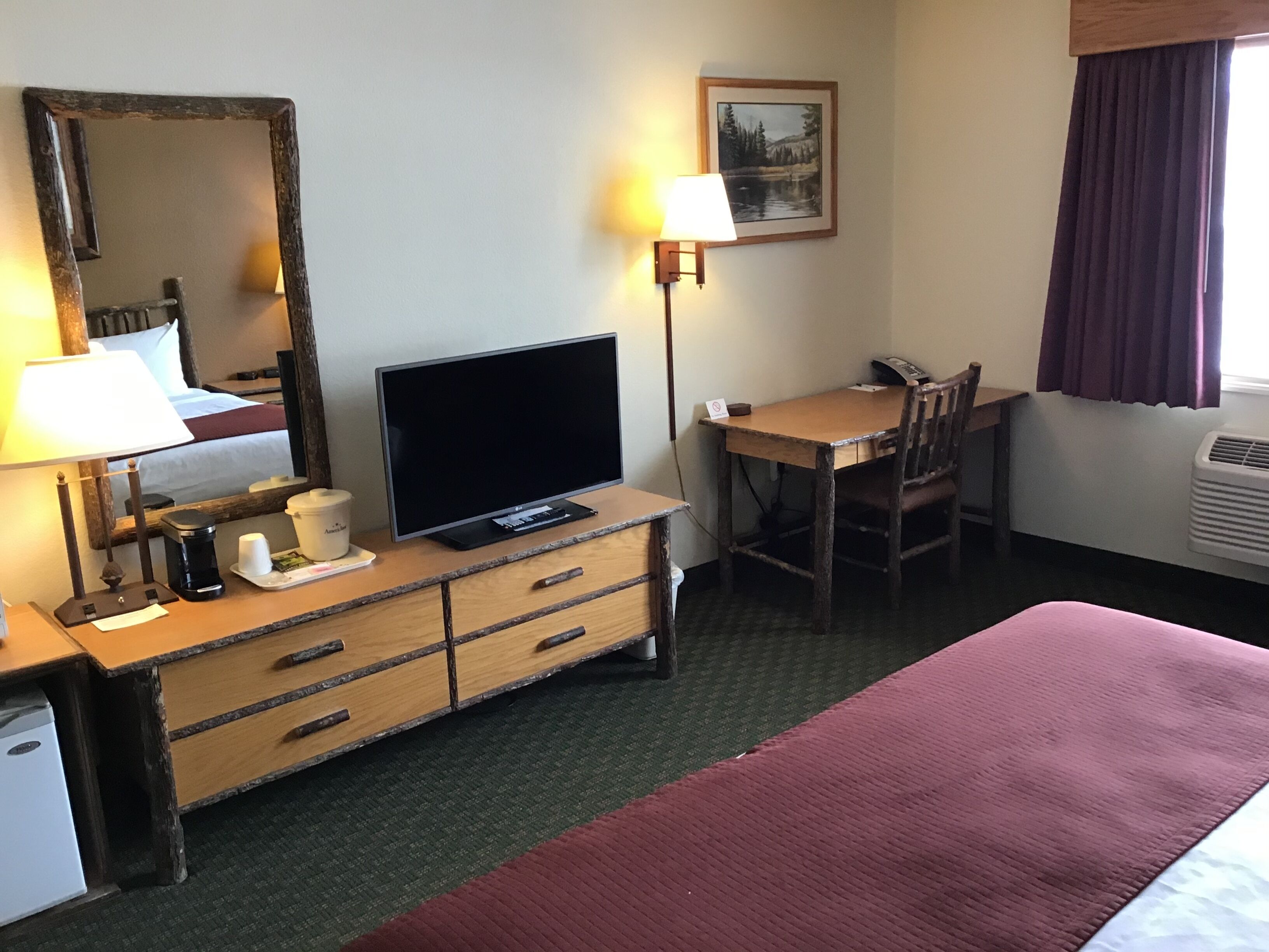 A hotel room with a bed, a desk with a chair, a television, a dresser, a lamp, and a painting on the wall.
