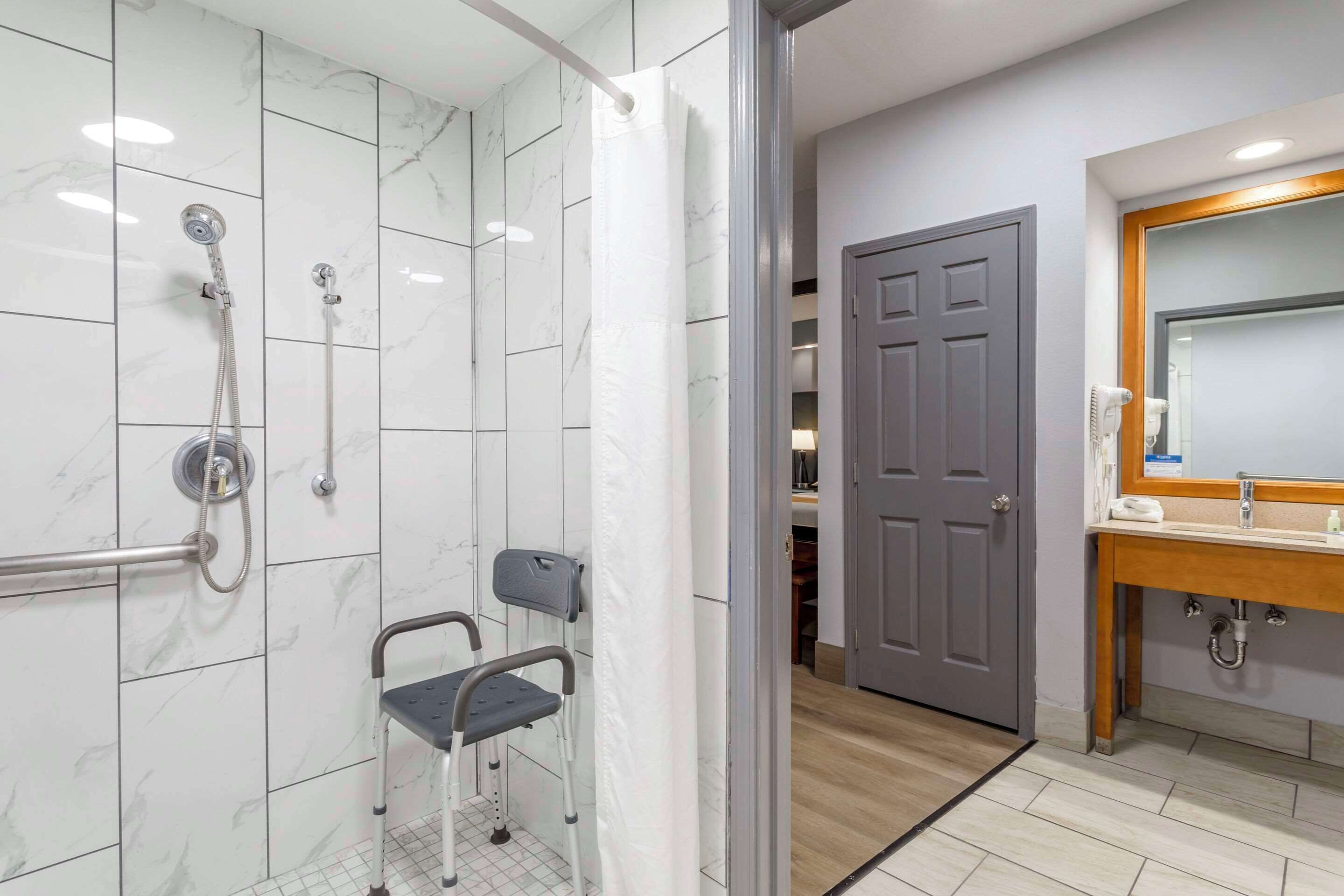 Un baño con ducha, una silla accesible para sillas de ruedas, un lavabo y una puerta que da a otra habitación.