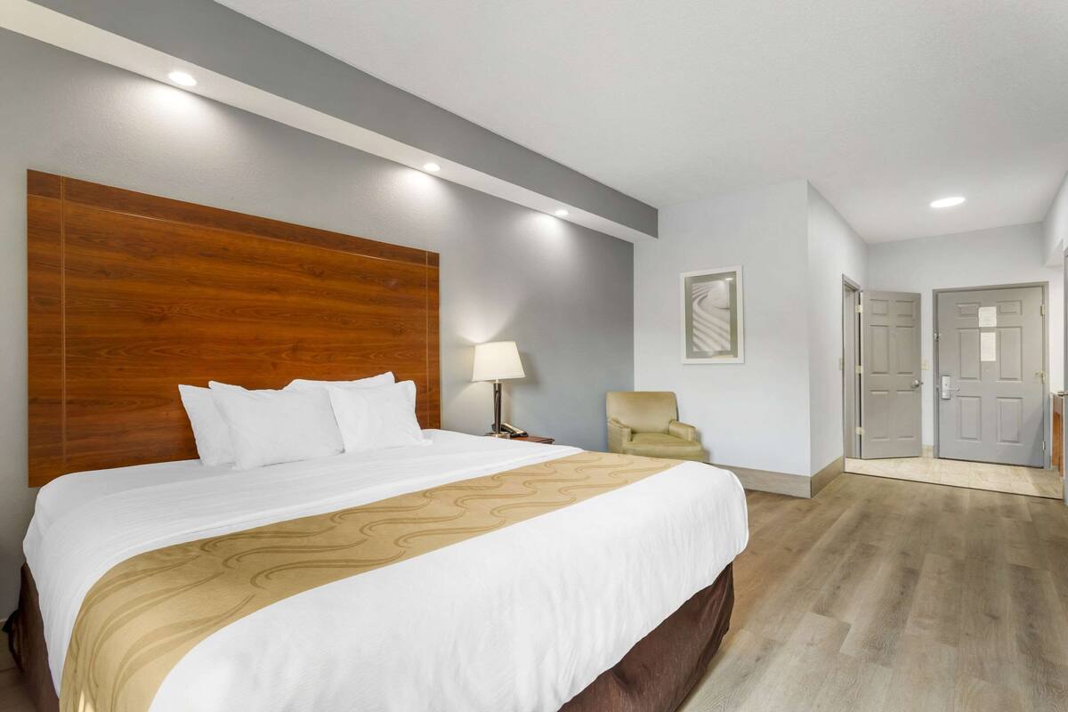 A hotel room with a large bed, a wooden headboard, a chair, and a door.